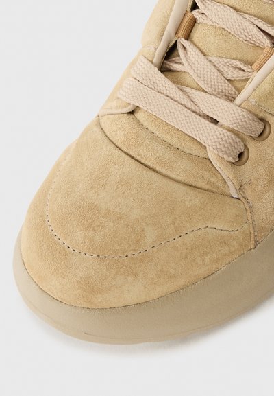 Beige suede sneakers feature a rounded toe, prominent stitching, and flat laces. The sole is a smooth, light tan material.