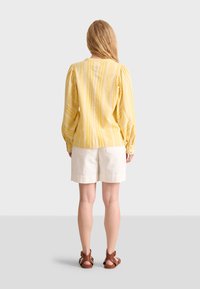 Blusa gialla a righe con maniche a sbuffo, abbinata a pantaloni corti color crema. La vista posteriore mostra una sottile texture e una vestibilità ampia. Sandali marroni completano il look.