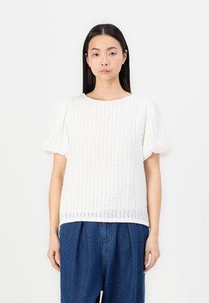White short-sleeve top with a textured ribbed design. Rounded neckline, loose fit, and soft fabric. Paired with blue denim trousers.