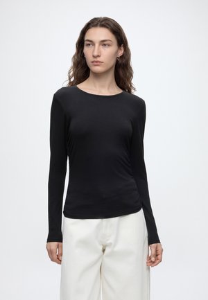 Femme aux longs cheveux ondulés portant un haut noir ajusté à manches longues et un pantalon blanc fluide, regardant légèrement sur le côté sur un fond uni.