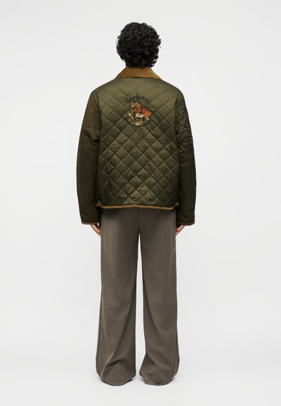 Άτομο με σγουρά μαλλιά που φοράει μια καπιτονέ μπουφάν ελιάς πράσινη με κεντημένο έμβλημα και καφέ γιακά, σε συνδυασμό με φαρδιά γκρι παντελόνια.