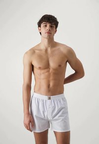 Boxer shorts bianchi in cotone con cintura elastica e piccolo bottone frontale. Texture liscia, design fino al ginocchio, indossati da un modello maschile a torso nudo.