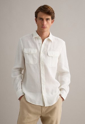 Giovane uomo con capelli castani indossa una camicia bianca a maniche lunghe con due tasche sul petto, pantaloni beige, in piedi davanti a uno sfondo semplice.