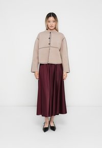 Giacca beige corta con cuciture e bottoni neri, abbinata a una lunga gonna di raso bordeaux. Scarpe nere basse completano l'outfit.