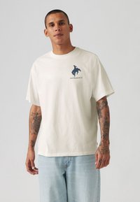 Cremefarvet bomulds t-shirt med korte ærmer, prydet med en marineblå logoillustration på venstre bryst og en afslappet, kasseformet pasform.