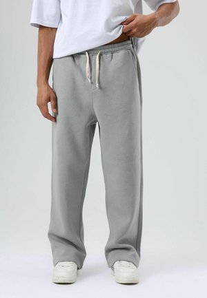 Grijze joggingbroek met een elastische tailleband en aantrekkoord. Zachte katoenen stof, wijd broekontwerp en zijzakken. Witte sneakers zichtbaar.