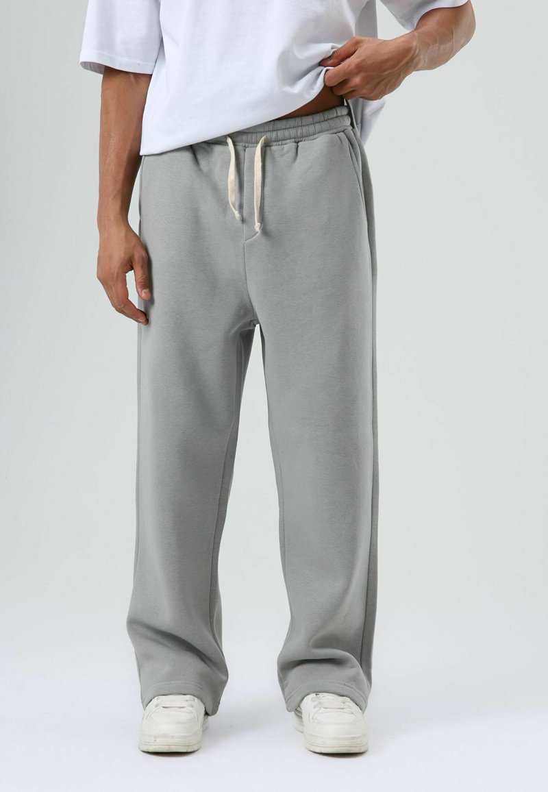 Pantalon de survêtement gris avec une taille élastique et un cordon de serrage. Tissu en coton doux, design à jambes larges, avec poches latérales. Baskets blanches visibles.