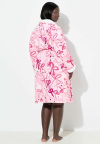 Růžový fleecový župan s opakujícím se vzorem mašlí v tmavší růžové a černé barvě. Má měkký bílý límec a dlouhé rukávy. Design s otevřeným zády.
