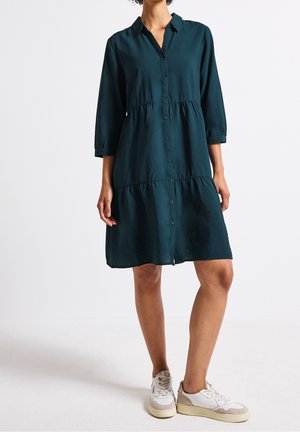 Kobieta ubrana w turkusową sukienkę z guzikami, warstwową, z rękawami do połowy przedramienia i białe trampki, stojąca na tle jednolitej białej ściany.