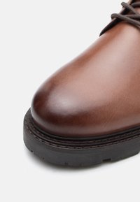 Καφέ δερμάτινο παπούτσι με στρογγυλή μύτη, λείο φινίρισμα και αντίθεση με μαύρη καουτσούκ σόλα. Διαθέτει καφέ κορδόνια που περνούν από τα μάτια.