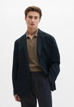 Man met lichtbruin haar, gekleed in een donkerblauw colbert, een olijfgroen overhemd met kraag en een donkere broek tegen een effen witte achtergrond.