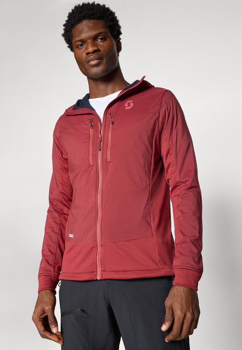 Man in een rode ritsjack met borstzakken en een witte t-shirt eronder, staande voor een vlakke lichte achtergrond.