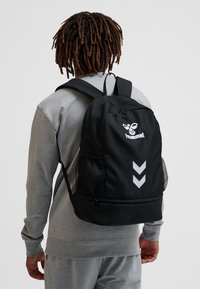 Persoon die een grijze sweater en broek draagt met een zwarte Hummel-rugzak met logo en twee witte chevrons aan de achterkant.