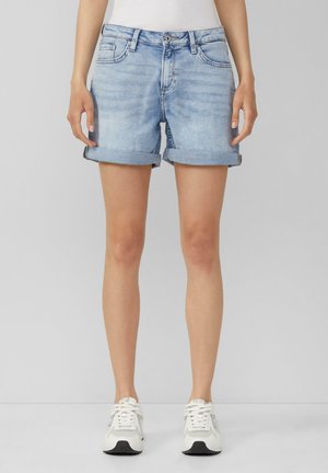 Mujer con pantalones cortos de mezclilla azul claro con los dobladillos enrollados y zapatillas blancas, de pie contra un fondo gris liso.