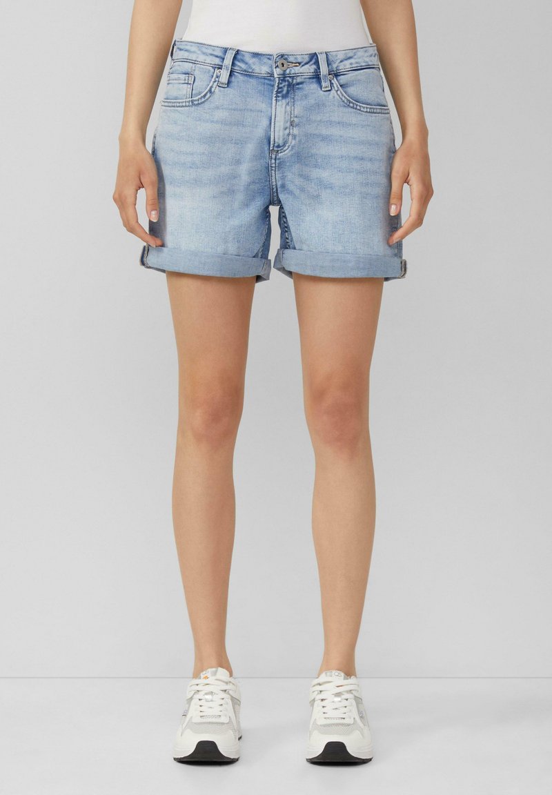 Femme portant un short en jean bleu clair retroussé et des baskets blanches, debout devant un fond gris uni.