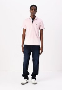 Lichtroze polo shirt met een marineblauwe kraag en bies, gecombineerd met donkerblauwe jeans en witte sneakers met zwarte accenten.