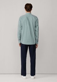 Hombre de pie de espaldas, vistiendo una camisa a cuadros verde y blanca, pantalones oscuros azules y zapatillas blancas, con un fondo liso.