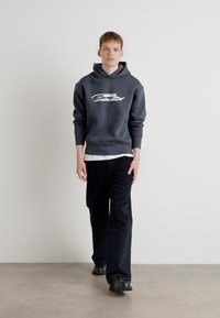 Gray hoodie with logo, white accents, relaxed fit, paired with black pants. Model stands on a smooth floor against a plain background.