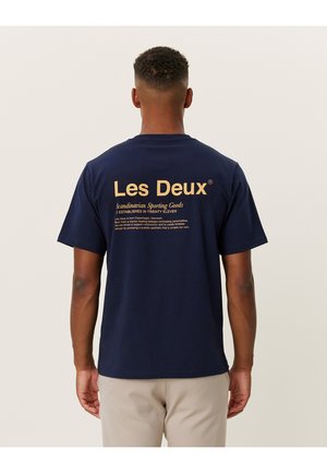 Tumedast sinise puuvilla T-särk, millel on suur beež tekst selja taga, sealhulgas brändi nimi "Les Deux" ja kirjeldav slogan.