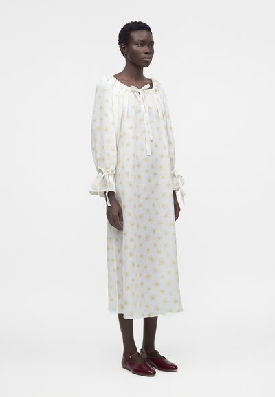 Γυναίκα που φοράει λευκό midi φόρεμα με κίτρινα λουλουδάτα σχέδια, μακριά φουσκωτά μανίκια δεμένα στους καρπούς, συνδυασμένο με μπορντό φλατ παπούτσια.