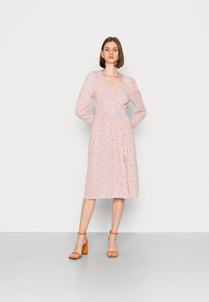 Vestido floral em rosa claro com detalhes em laranja, apresentando decote em V, mangas longas, cintura elástica e saia de comprimento médio. Usado com saltos laranjas.