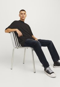 Camiseta negra de gran tamaño, jeans de mezclilla oscuros y zapatillas negras con cordones blancos. Sentado en una silla de metal plateada con listones verticales.