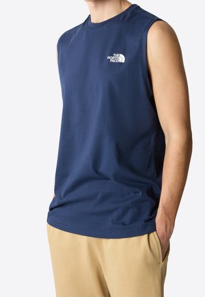 Mężczyzna w granatowej koszulce bez rękawów z logo The North Face na piersi i beżowych spodniach, z rękami w kieszeniach.