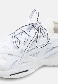 White athletic shoe with mesh upper, rubber sole, black accents, and unique cutouts. Features flat laces and embossed logo detail.