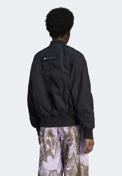 Fekete bomber dzseki fényes anyagból, bordázott mandzsettákkal és aljjal rendelkezik. A hátsó részén egy márkás címke látható, mintás nadrággal párosítva.