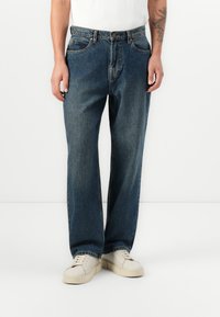 Jeans de mezclilla azul oscuro con un corte relajado, diseño de cinco bolsillos y cierre de botón. Combinados con zapatillas blancas, mostrando una estética simple.