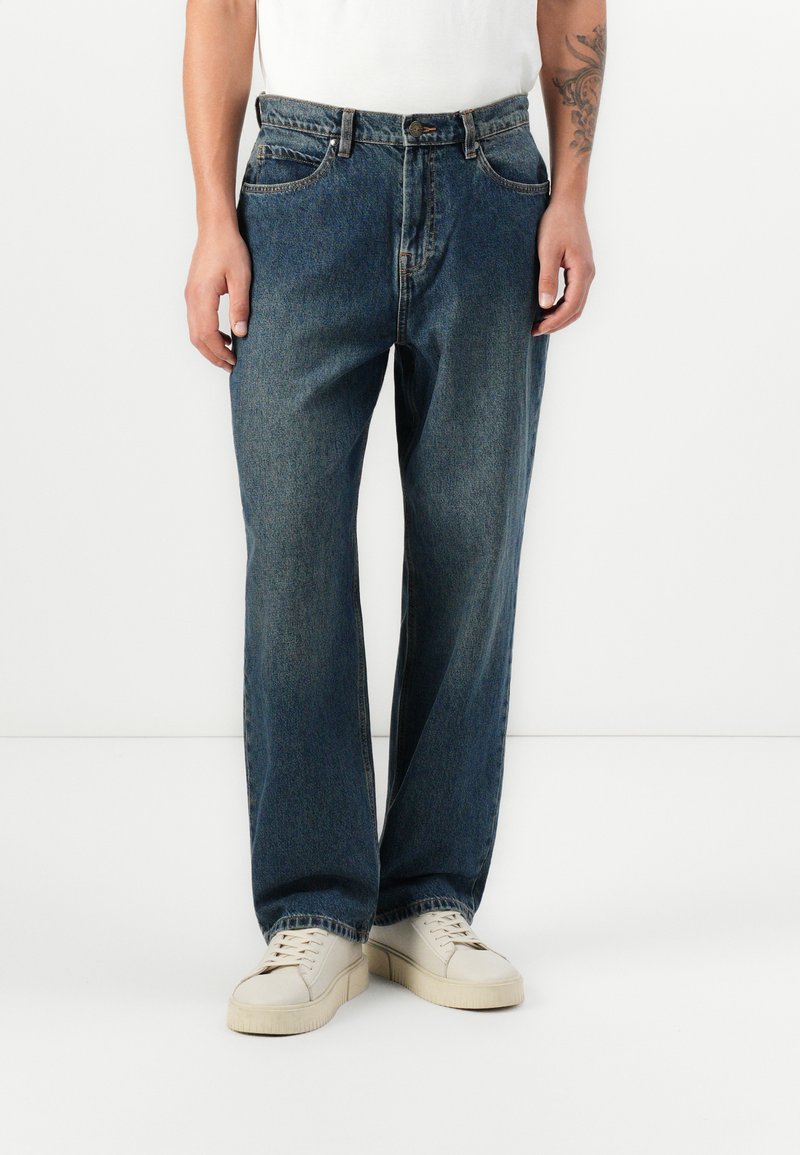 Jeans de mezclilla azul oscuro con un corte relajado, diseño de cinco bolsillos y cierre de botón. Combinados con zapatillas blancas, mostrando una estética simple.
