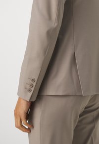 Blazer ajusté beige clair avec une texture lisse. Il est doté d'une fente arrière et de cinq boutons aux poignets, offrant un look moderne et élégant.