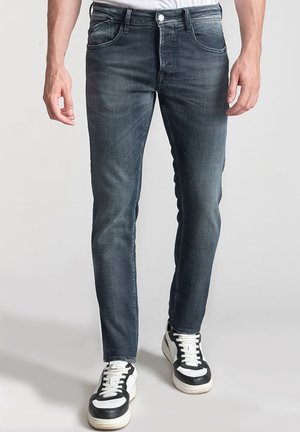 Jean en denim bleu foncé avec une coupe slim, cinq poches, un léger délavage et une couture contrastée, associés à des baskets noires et blanches.