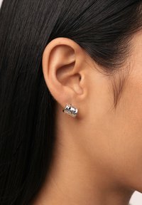 Pendiente de aro de metal plateado con superficie texturizada, que presenta formas curvas y un acabado pulido, llevado en la oreja de un modelo.