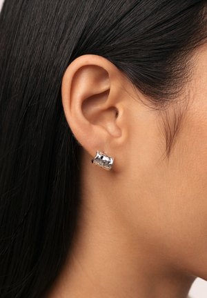 Boucle d'oreille en métal argenté avec une surface texturée, présentant des formes cintrées et une finition polie, portée sur l'oreille d'un modèle.