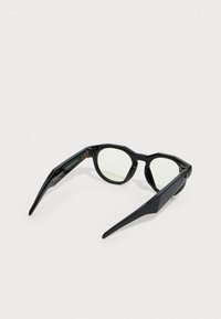 Mustat aurinkolasit, joissa on kulmikas muotoilu, vihreät linssit ja geometriset yksityiskohdat. Sankojen pinta on kiiltävä ja niissä on erottuvat templet.