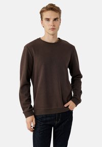 Sudadera marrón de manga larga con textura acanalada, cuello redondo y corte relajado. Combinada con jeans de mezclilla oscuros. Diseño minimalista.