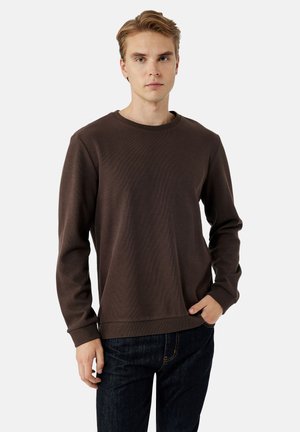 Sudadera marrón de manga larga con textura acanalada, cuello redondo y corte relajado. Combinada con jeans de mezclilla oscuros. Diseño minimalista.