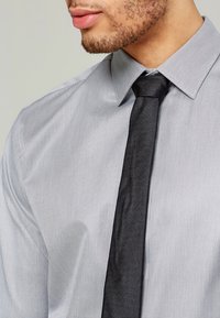 Grå rutiga skjortan med en strukturerad krage, matchad med en texturerad svart slips. Skjorttyget verkar vara slätt och lätt. 