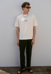 Camiseta corta de manga con diseño bordado, combinada con pantalones negros y sandalias negras de tipo slide. Los accesorios incluyen un collar de cadena de plata.
