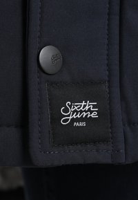 Mörkblå tygjacka med en slät yta. Har en svart knapp och en etikett med vit skrift som läser "Sixth June Paris."