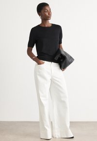Black short-sleeved knit top paired with high-waisted white wide-leg trousers. Model holds a black leather clutch. Smooth texture, modern design.