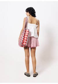 Rode en roze geweven tas met een geometrisch patroon, over de schouder gedragen. Model in een witte top en roze gestreepte shorts, staande tegen een witte achtergrond.