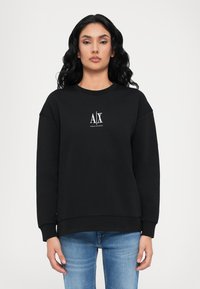 Juodas sweatshirt su apvalia iškirpte, nukritusiais pečiais ir baltu logotipu ant krūtinės. Pagamintas iš minkštos, tekstūruotos medžiagos. Derinamas su mėlynais džinsais.