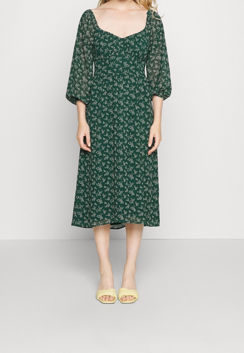 Robe midi à fleurs vertes avec manches bouffantes transparentes et encolure carrée. Associée à des sandales à enfiler jaune clair.