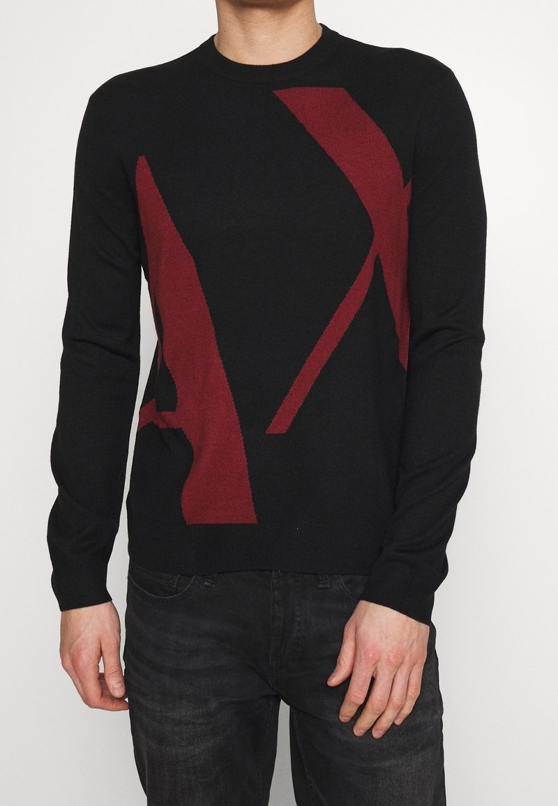 Homme portant un pull noir avec de grandes formes abstraites rouge foncé, associé à un jean foncé, sur un fond clair uni.
