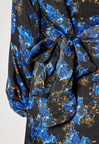 Tissu noir avec un motif floral bleu et jaune noué en nœud à la taille, montrant une manche longue avec poignet froncé.