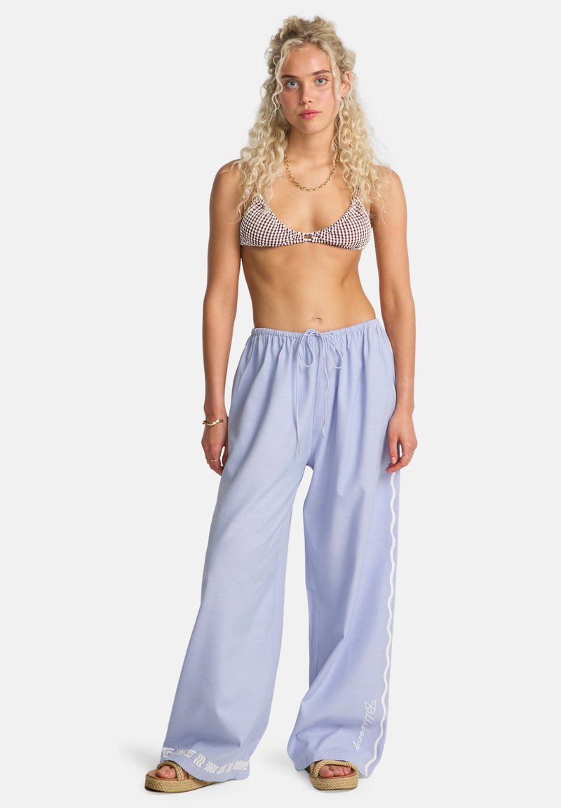 Hellblaue, weit geschnittene Hosen mit Kordelzug in der Taille und weißen Stickereien am Saum. Kombiniert mit einem braunen, karierten Bikini-Oberteil.