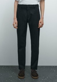 Pantaloni neri con taglio dritto, realizzati in tessuto texturizzato, con chiusura a bottoni e tasche frontali. Abbinati a scarpe marroni.