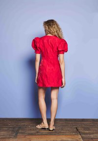 Γυναίκα με σγουρά ξανθά μαλλιά στέκεται μπροστά σε έναν μπλε τοίχο φορώντας ένα κοντό κόκκινο φόρεμα με φουσκωτά μανίκια και μπεζ σανδάλια με χαμηλό τακούνι.